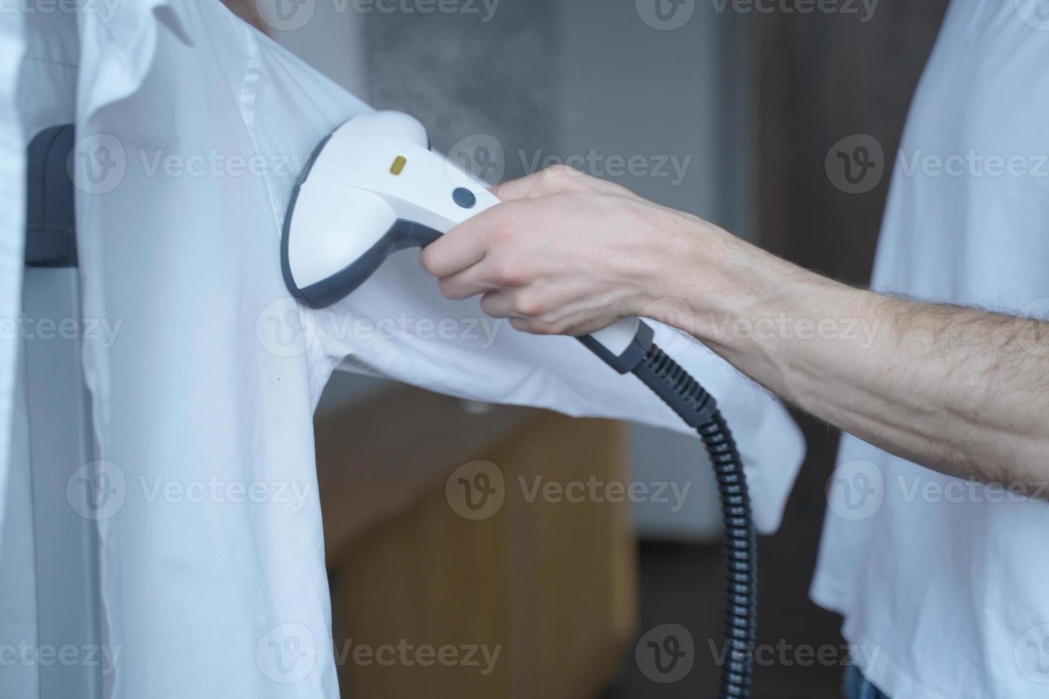 abgeschnittene Aufnahme eines Mannes, der einen elektrischen Kleiderdampfer benutzt, während er zu Hause ein klassisches Hemd dämpft foto