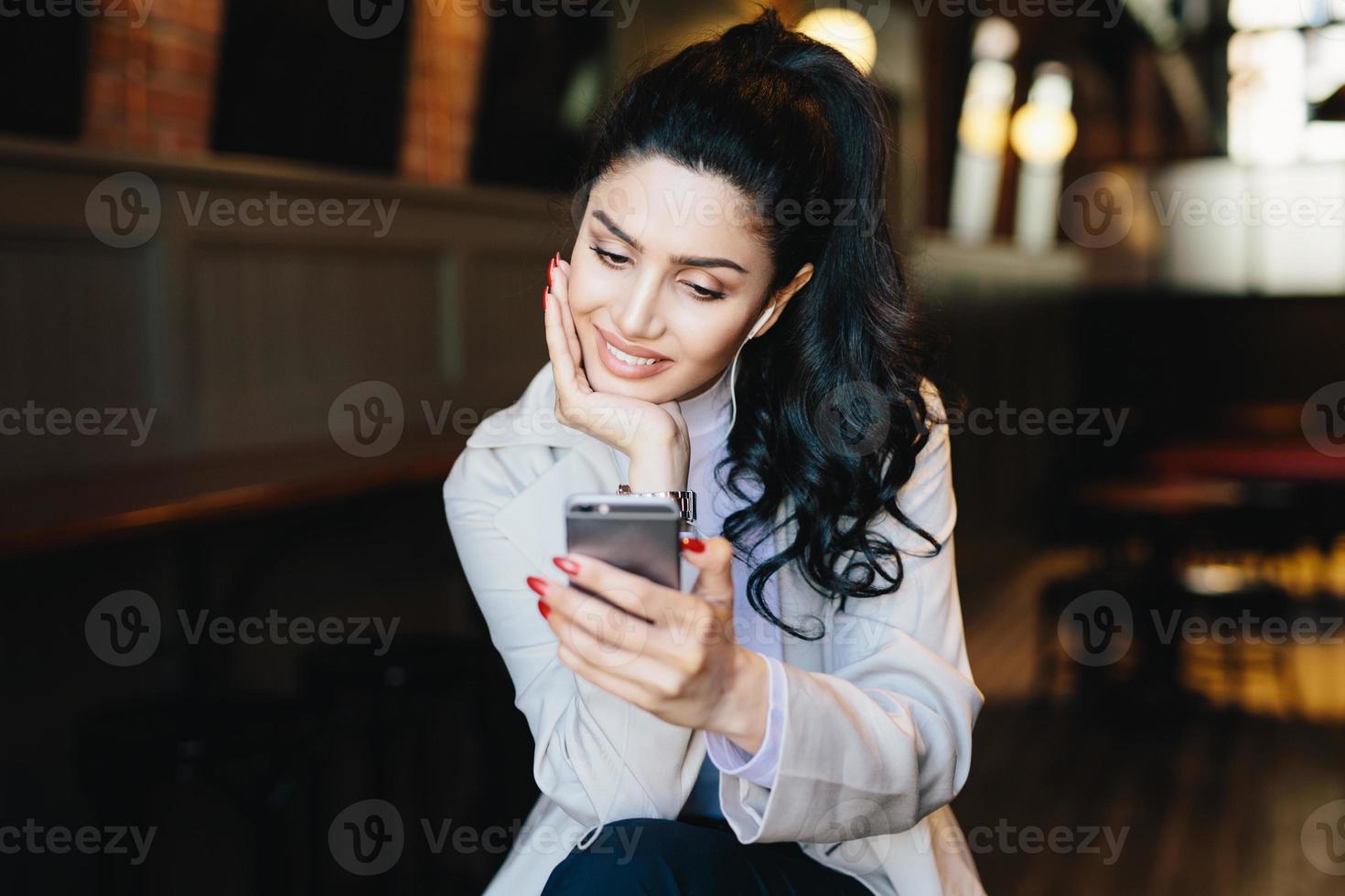 angenehm aussehende elegante frau mit dunklen haaren mit roten nägeln in weißem mantel, die lieder auf dem handy hören, e-mails mit elektronischen geräten abrufen, videoanrufe mit freunden tätigen foto