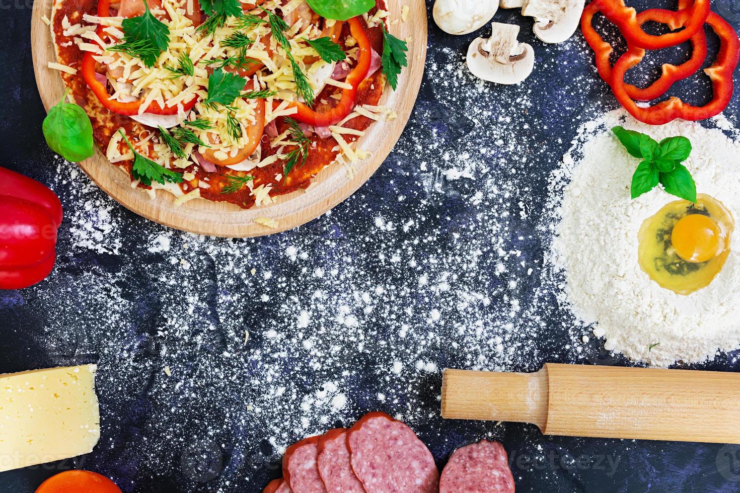 roher Pizzateig mit Zutaten und Gewürzen auf dunklem Hintergrund. Ansicht von oben foto