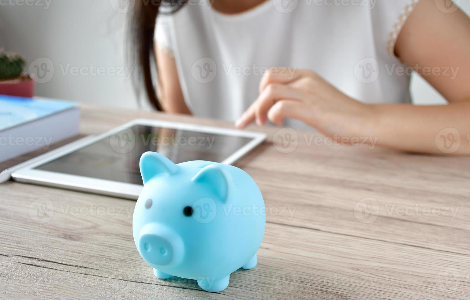 eine frau, die die einsparungen auf dem ipad berechnet. Auf dem Holztisch befindet sich auch ein blaues Sparschwein, um Geld zu sparen und das Haushaltseinkommen auf dem Tablet zu sparen. foto