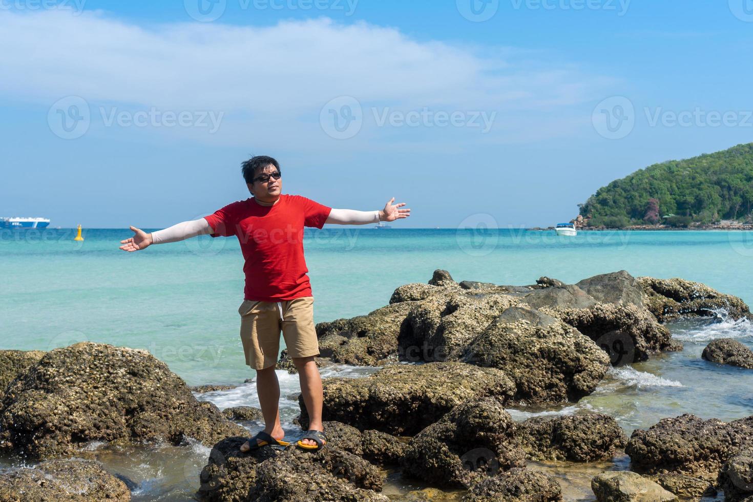 asiatischer mann trägt rotes t-shirt, das auf offenen händen des felsens steht, fühlt freiheit und entspannt sich an einem sonnigen tag am blauen seestrand foto