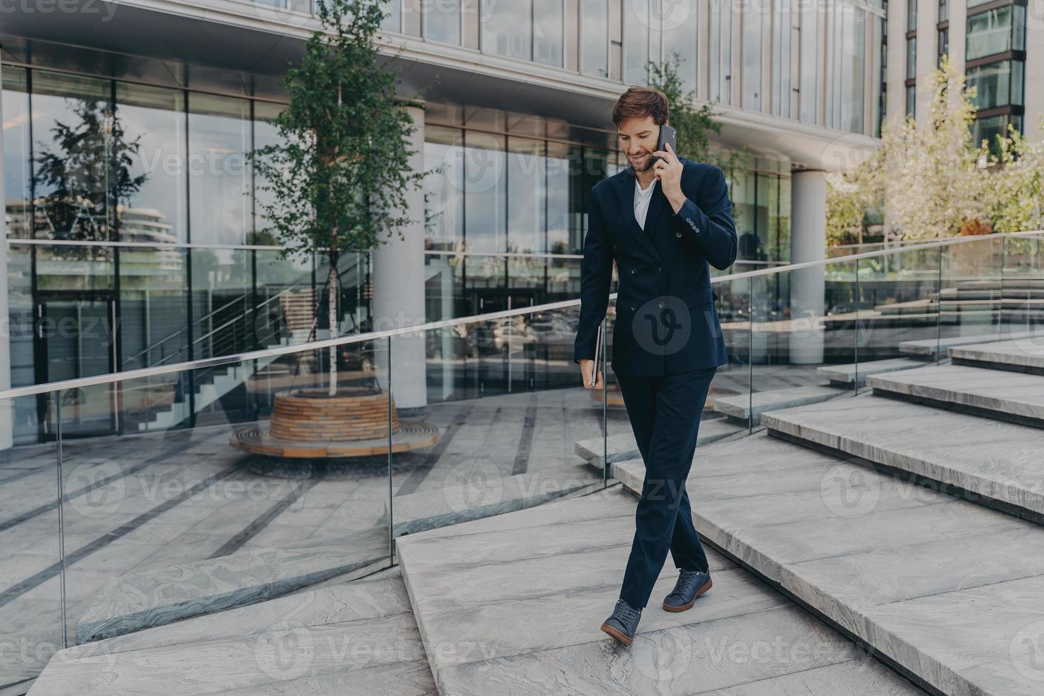 glücklicher kaukasischer geschäftsmann, der auf dem smartphone spricht, während er entlang des bürozentrums geht foto