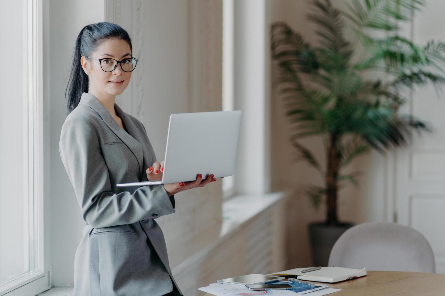 Verwaltungsfrau steht in der Nähe des Desktops, hält einen modernen Laptop-Computer in der Hand, nutzt eine drahtlose Verbindung zum Internet, trägt ein formelles graues Outfit, sucht nach Informationen auf der Webseite, arbeitet entfernt foto