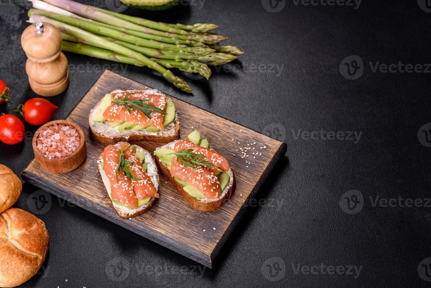 Toastsandwich mit Butter, Avocado und Lachs, dekoriert mit Rucola und Sesamsamen, auf schwarzem Steinhintergrund foto