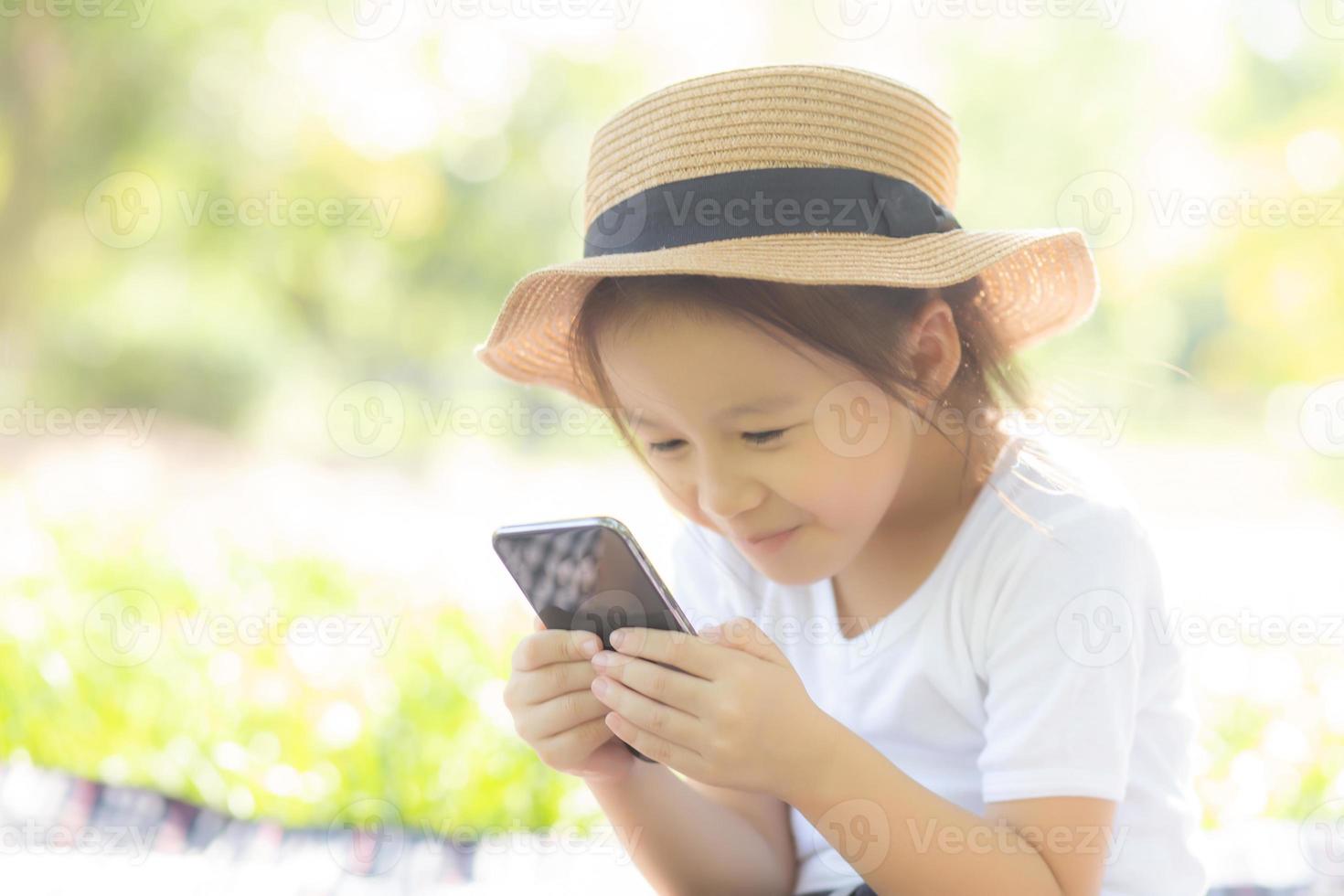 Schönes asiatisches Kind, das mit einem intelligenten Handy im Garten lächelt, Kind hat eine Leidenschaft, digitale Spiele auf dem Smartphone im Internet-Netzwerk im Park mit einem glücklichen, Lifestyle- und Technologiekonzept zu spielen. foto