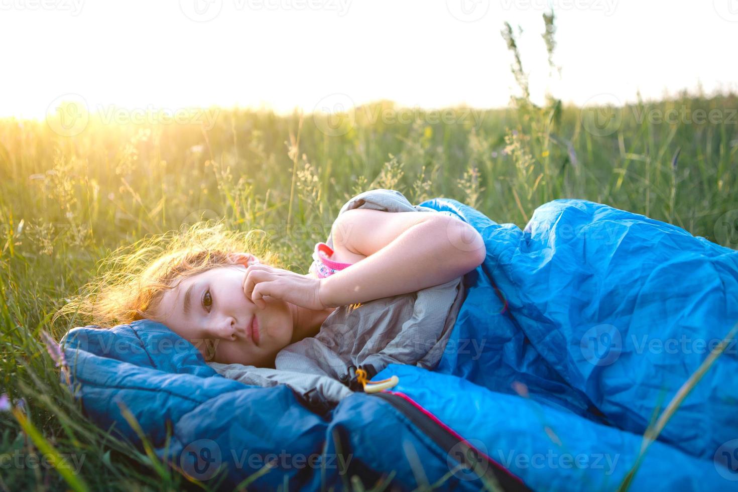 Das Mädchen ist unzufrieden mit kratzenden Mückenstichen, das Kind schläft bei einem Campingausflug in einem Schlafsack auf dem Gras. umweltfreundliche erholung im freien, sommerzeit. Schlafstörung, abweisend. foto
