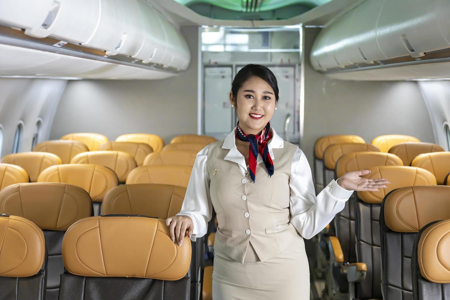 asiatische flugbegleiterin, die mit einem lächeln in der mitte des gangs im flugzeug posiert, um den passagier an bord mit sitz im hintergrund zu begrüßen foto