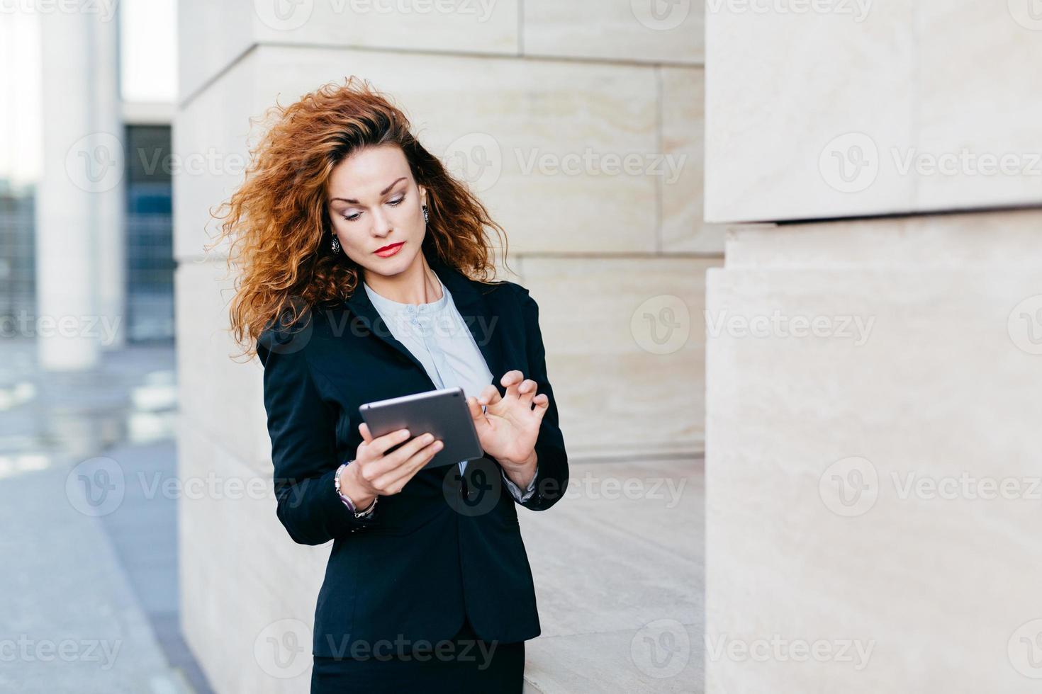 elegante Dame mit lockigem Haar, schwarzem Anzug, Nachrichten eingeben oder Geschäftsbericht erstellen, während sie einen Tablet-Computer verwendet. Unternehmerin, die an neuen Geschäftsprojekten arbeitet. Geschäft und Karriere foto
