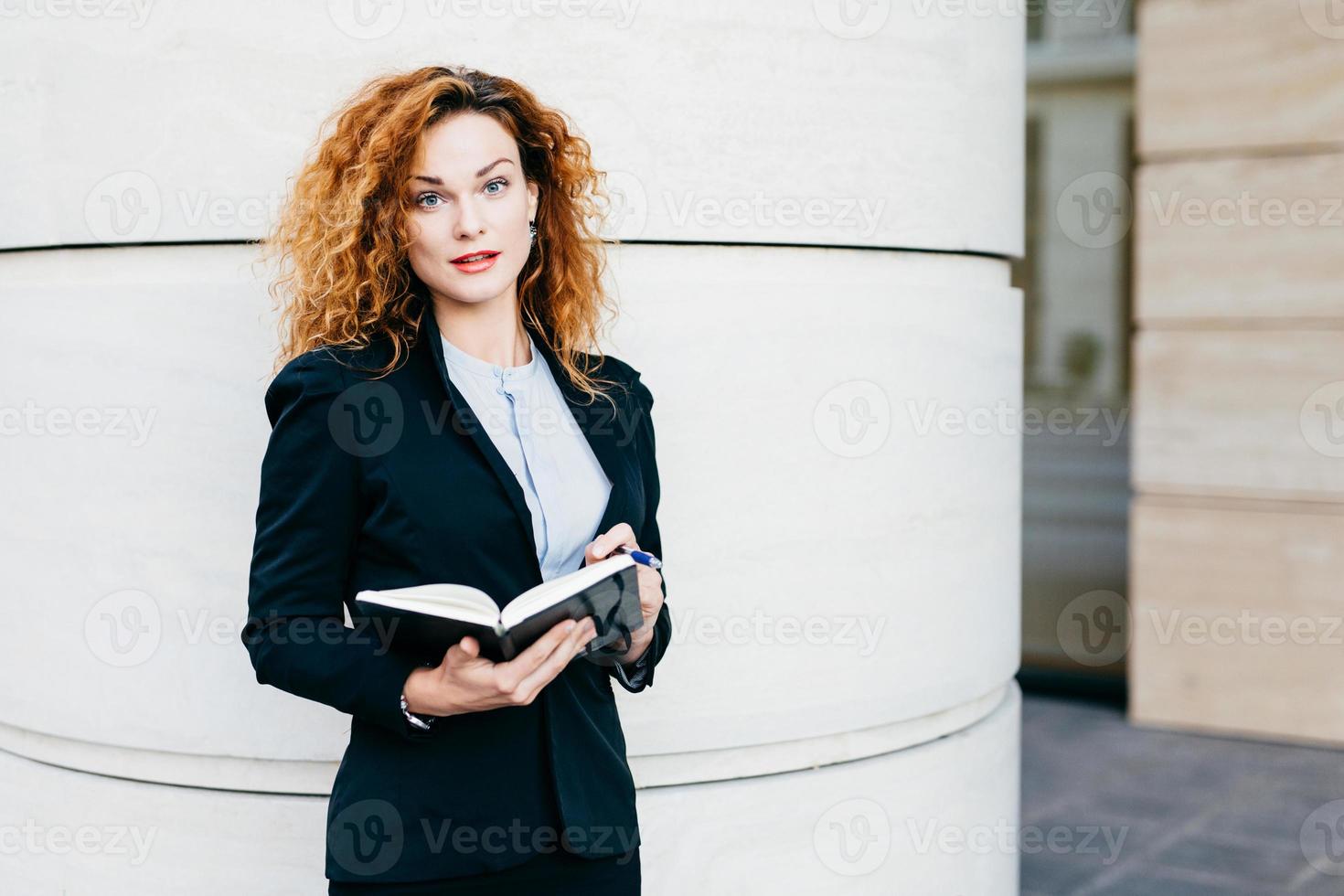 porträt einer geschäftsfrau mit lockigem haar, rot bemalten lippen, eleganter kleidung, die in ihr tagebuch schreibt. gutaussehendes damenmodell mit attraktivem aussehen, das ihr notizbuch mit stift hält foto