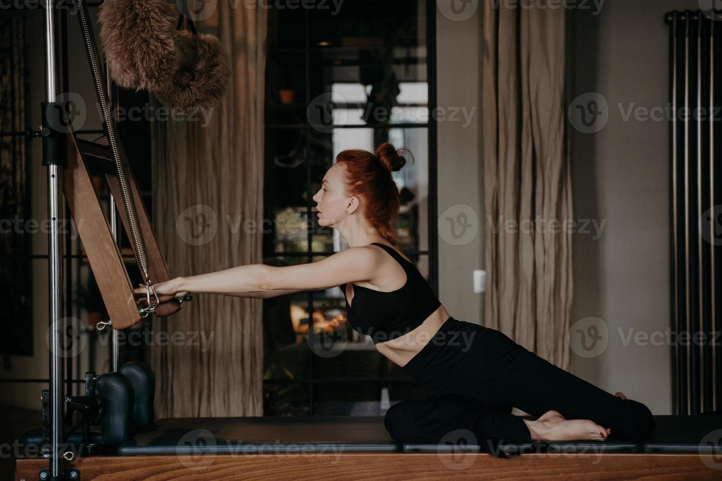 sportliche rothaarige frau, die während des pilates-trainings den athletischen körper auf dem cadillac reformer dehnt foto