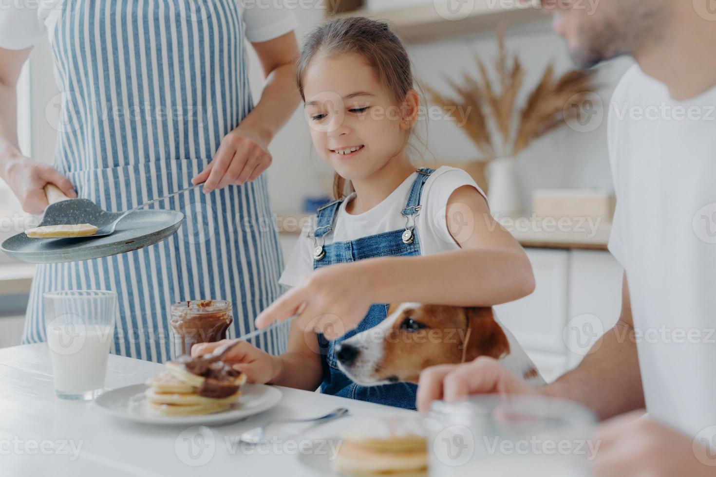 kleines kind, ihr vater und ihr hund sitzen zusammen am küchentisch, essen frisch zubereitete pfannkuchen, mutter in schürze steht in der nähe und hält pfanne. familie appetitlich leckere desset in der küche. kochen, ernährungskonzept foto