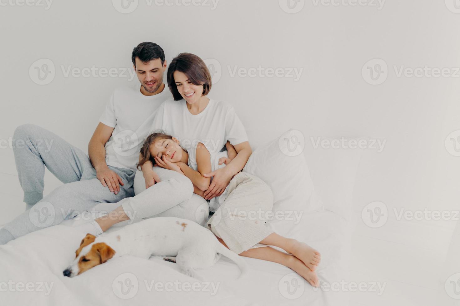 Foto der schönen Familie verbringen Freizeit zusammen, bleiben lange im Bett, haben gesund geschlafen, Rassehund liegt in der Nähe der Besitzer, genießt Komfort und Entspannung, Leerzeichen auf hellem Hintergrund für Promo