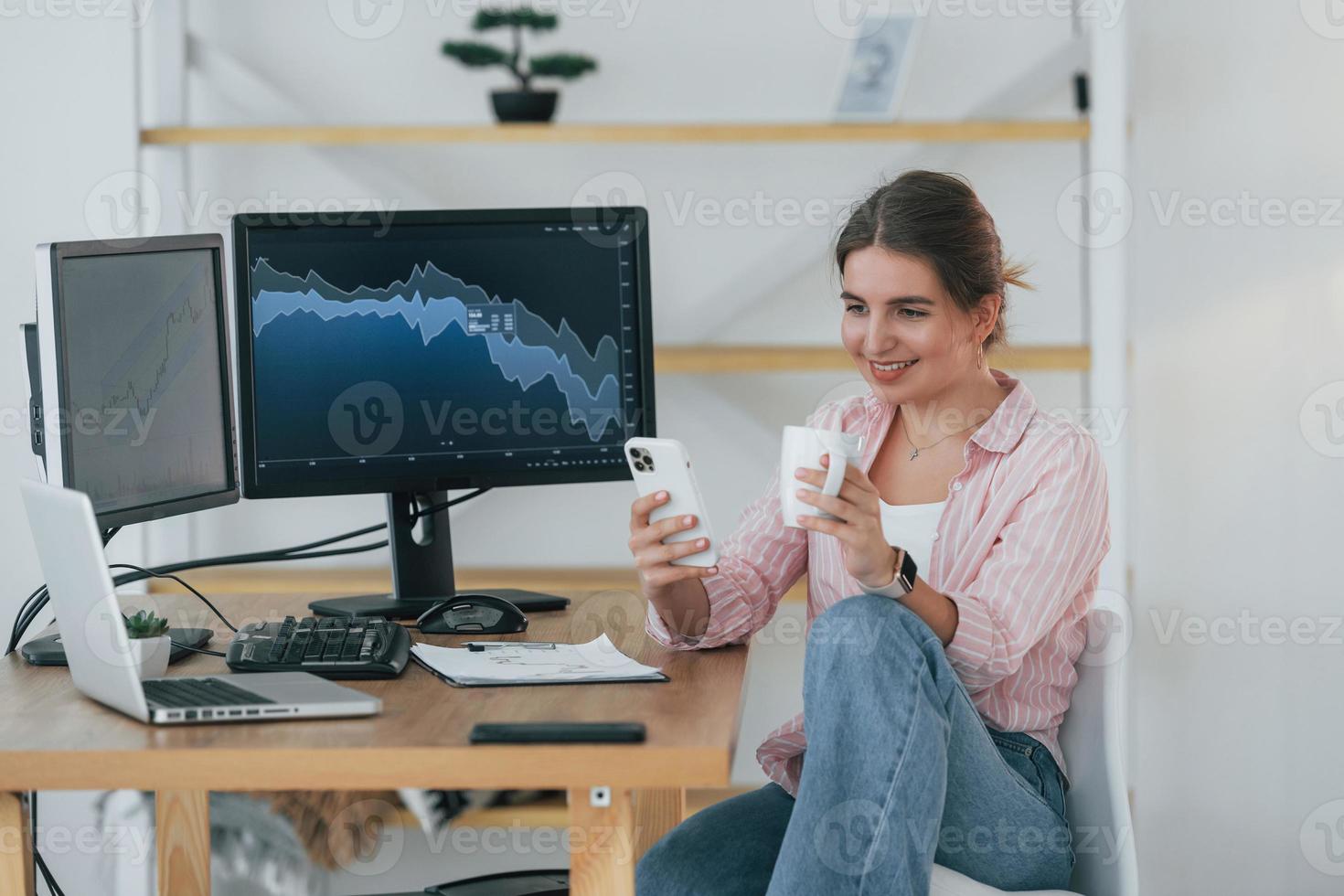 Sitzen mit Tasse Getränk und Smartphone in den Händen. weibliche börsenmaklerin in freizeitkleidung arbeitet im büro per pc foto