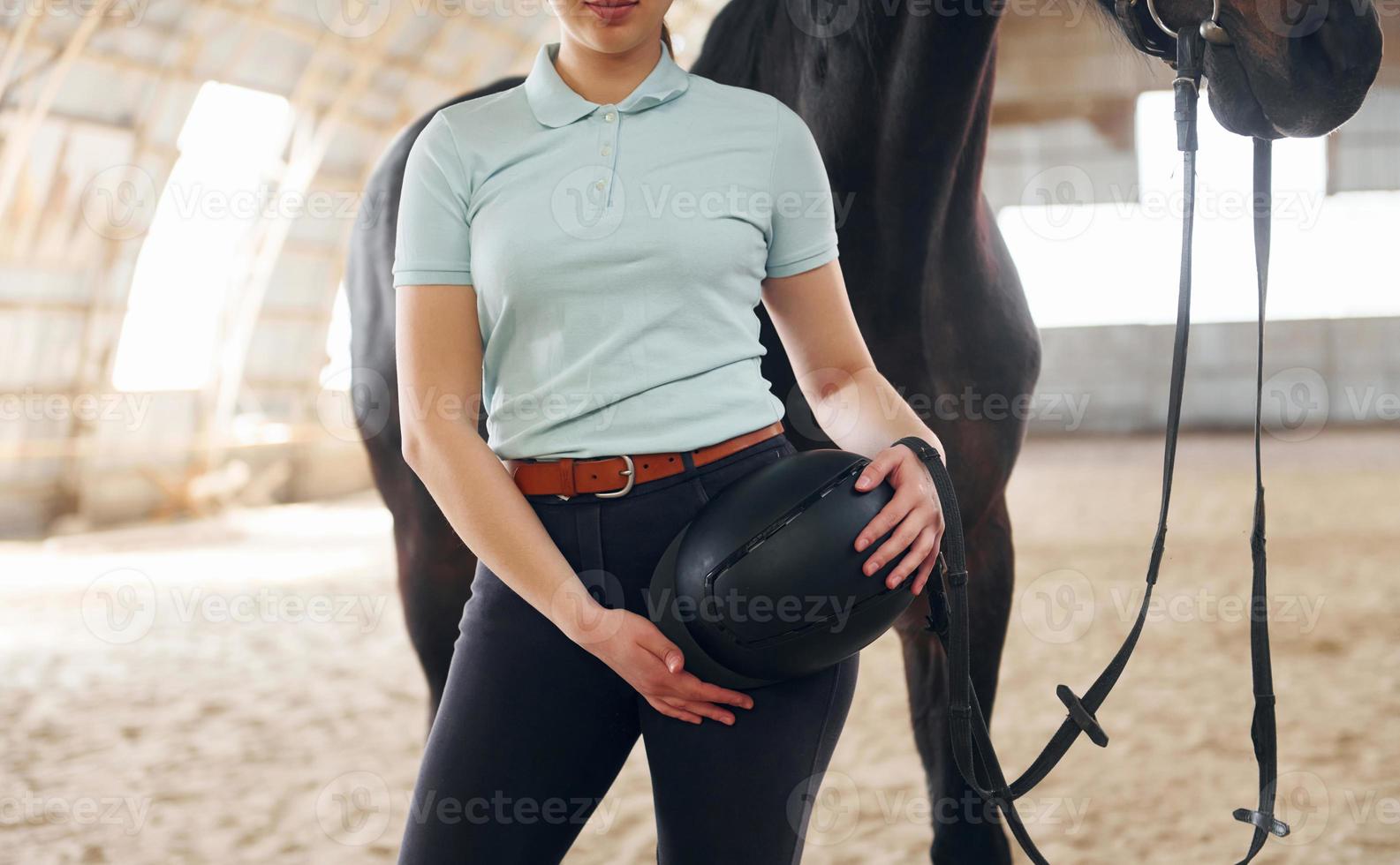 Stehen und Schutzhut in den Händen halten. eine junge frau in jockeykleidung bereitet sich auf einen ritt mit einem pferd auf einem stall vor foto