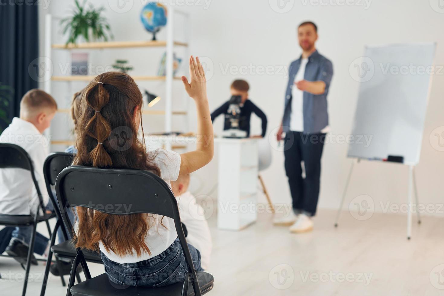 Junge mit Mikroskop, Mädchen hebt Hand hoch. gruppe von kinderschülern im unterricht in der schule mit lehrer foto