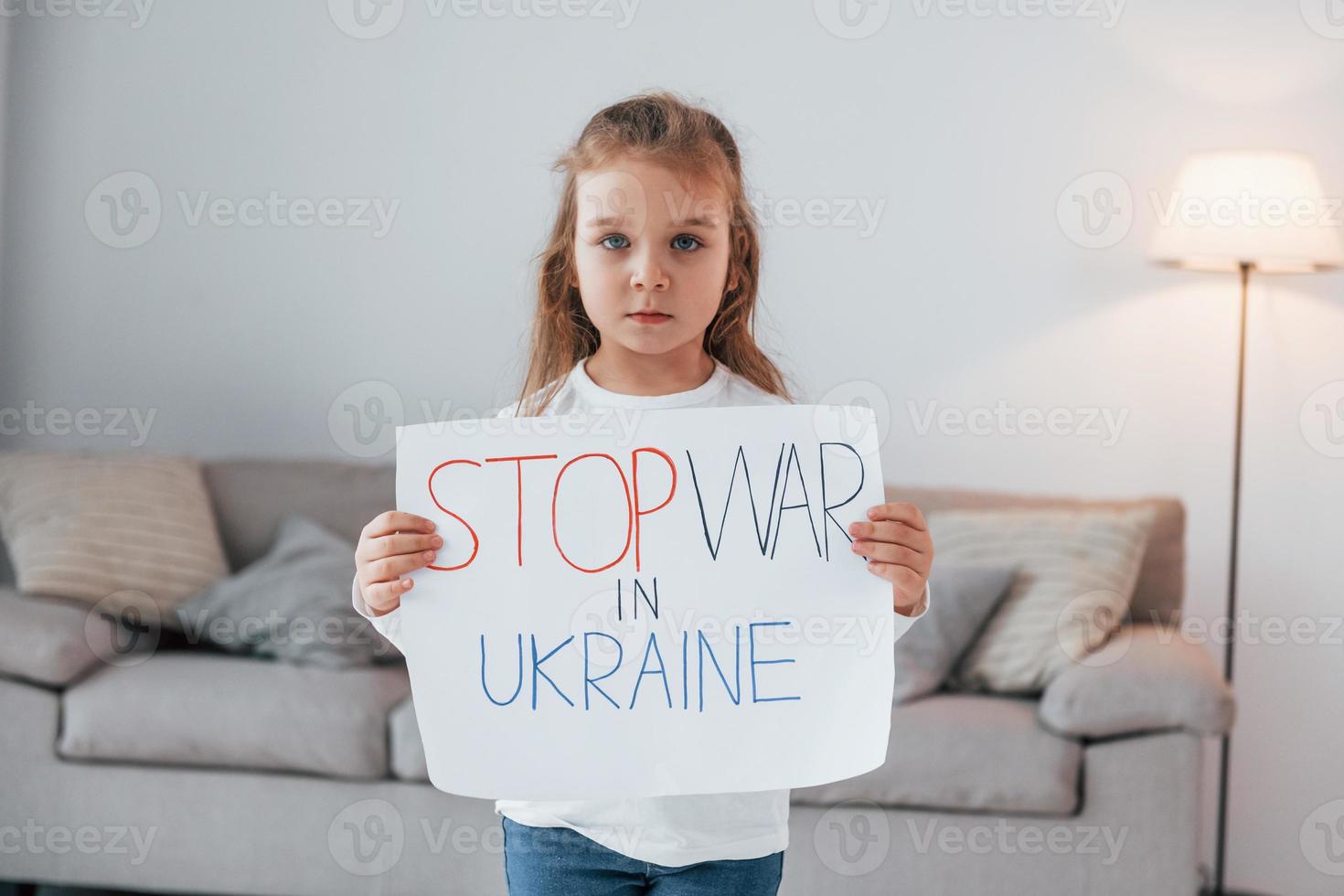 kleines mädchen, das zu hause steht und banner mit stop war im ukrainischen text hält foto