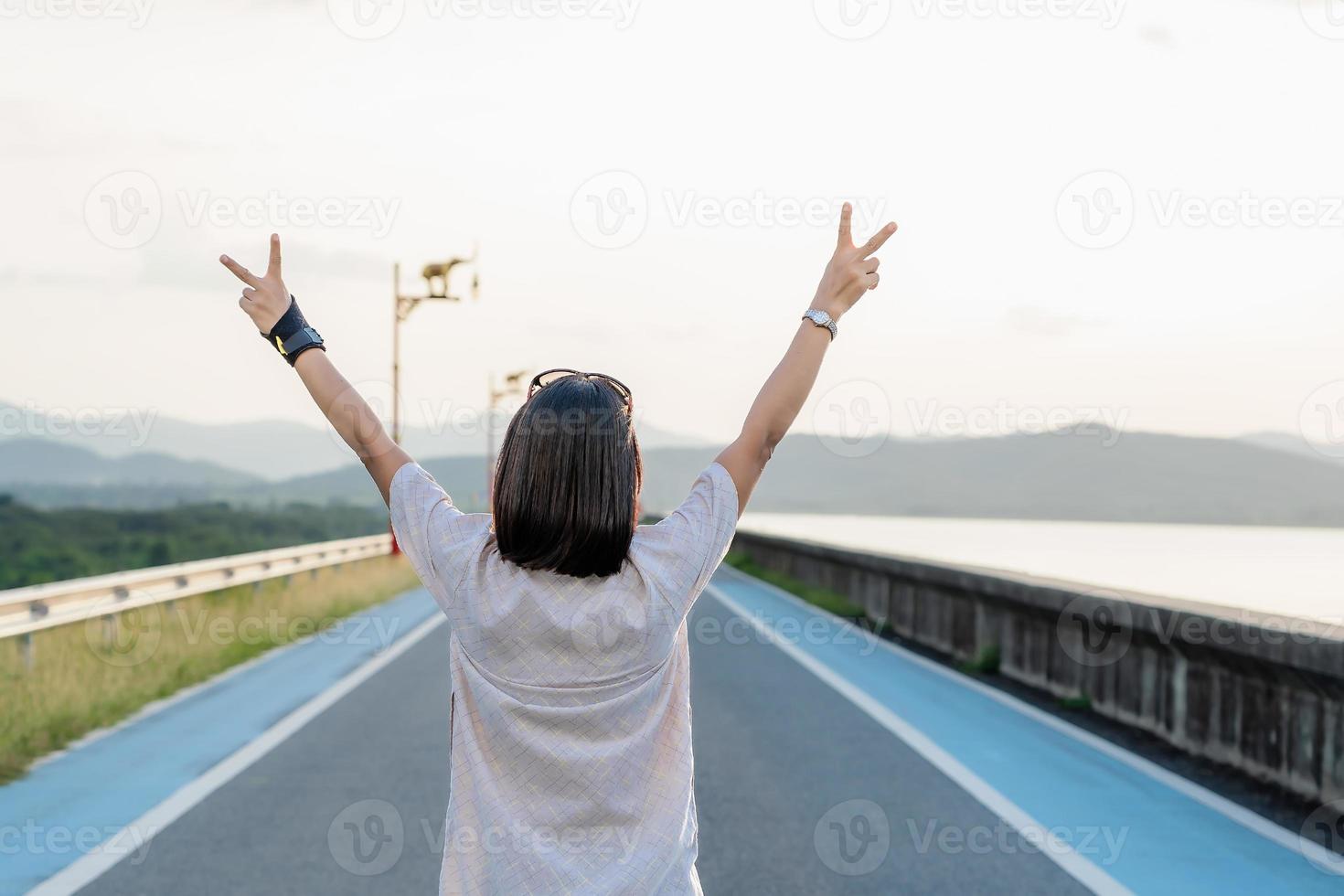 Rückansicht einer Touristin, die die Hände in die Luft hebt. Reisender, der auf einer Urlaubsreise auf der Straße des Damms mit einem Sonnenuntergang im Hintergrund steht. asiatische dame, die die natur im urlaub genießt. Freiheitskonzept foto