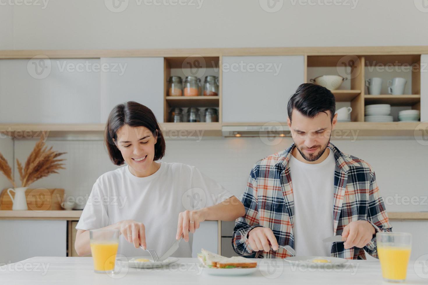 familie ehepaar posieren am küchentisch, lecker frühstücken, über den tag sprechen, spiegeleier und burger essen, frischen apfelsaft trinken, lässig gekleidet, häusliche atmosphäre genießen foto