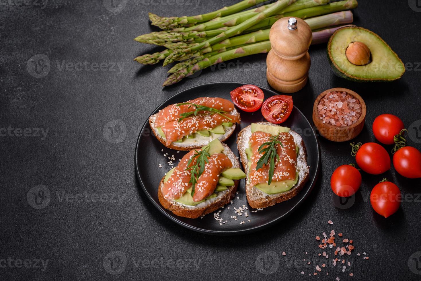 Toastsandwich mit Butter, Avocado und Lachs, dekoriert mit Rucola und Sesamsamen, auf schwarzem Steinhintergrund foto