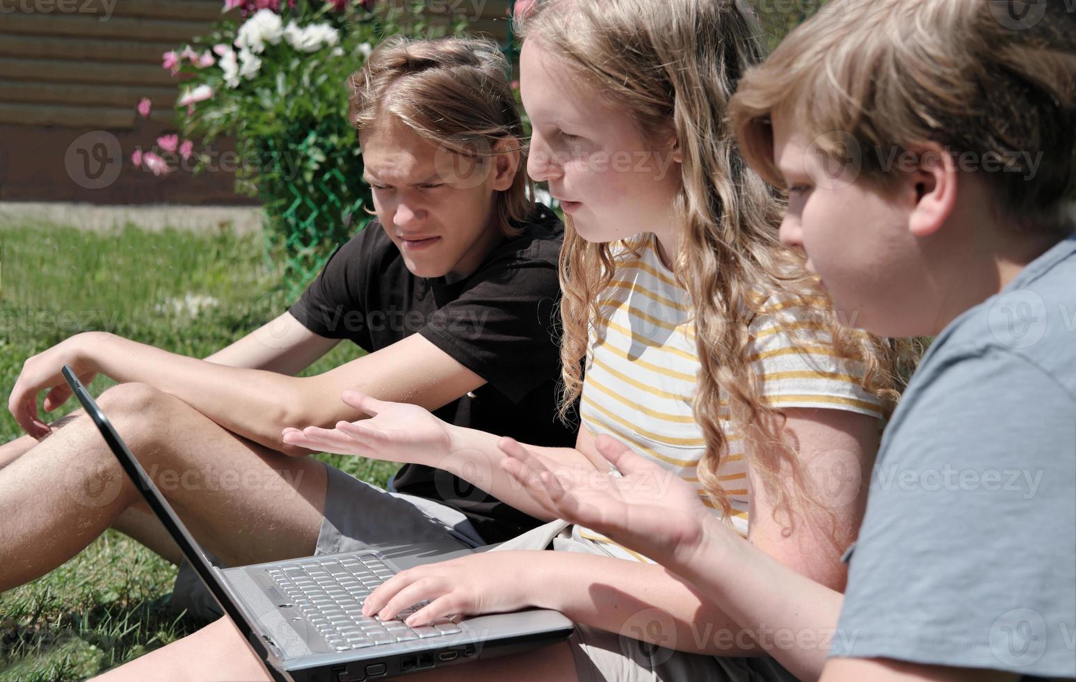 Zwei Teenager und ein Mädchen sitzen auf einer Wiese und unterhalten sich per Laptop per Video. kommunikations- und zusammengehörigkeitskonzept. emotionale Teenager foto
