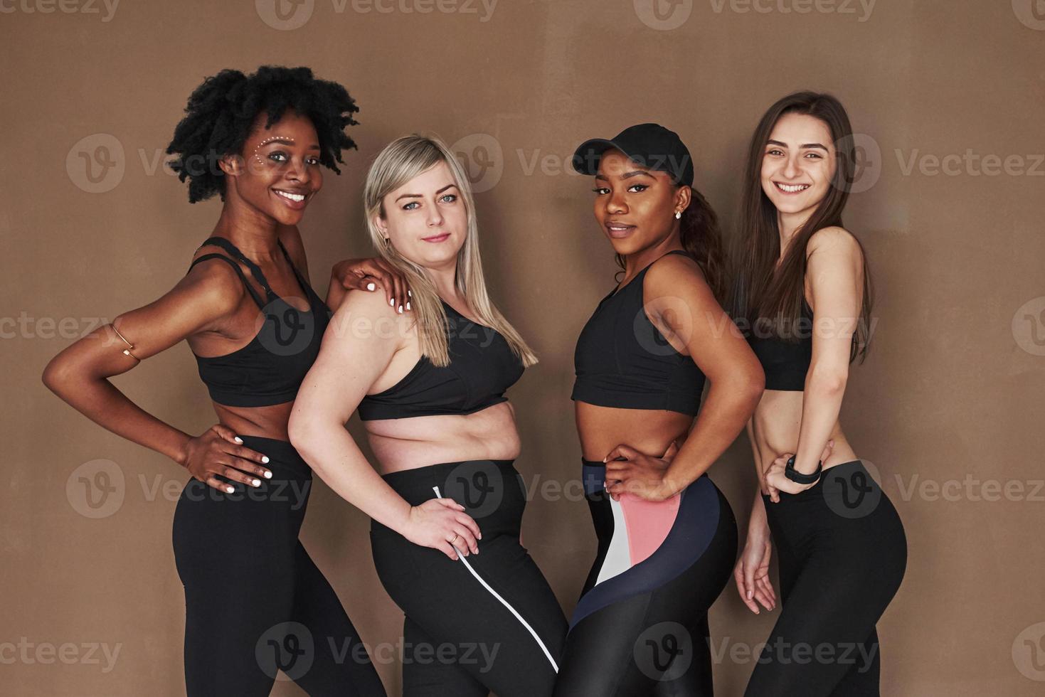 Hände auf die Hüften. Gruppe multiethnischer Frauen, die im Studio vor braunem Hintergrund stehen foto