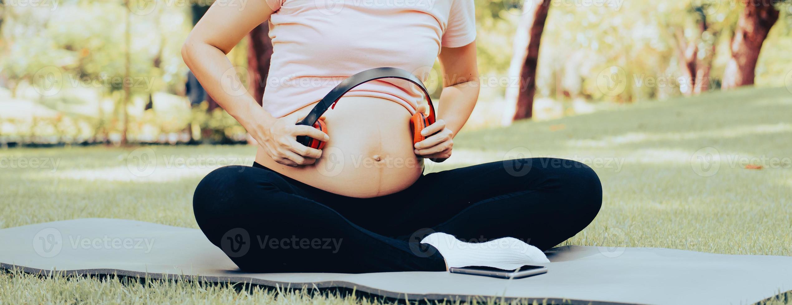 Schwangere Frau, die draußen im Park sitzt und ihren dicken Bauch mit roten Kopfhörern berührt, lässt ihr Baby beruhigende Harmoniemusik hören. schwangerschaftsreise, mutterschafts- und fötusentwicklungskonzept. foto