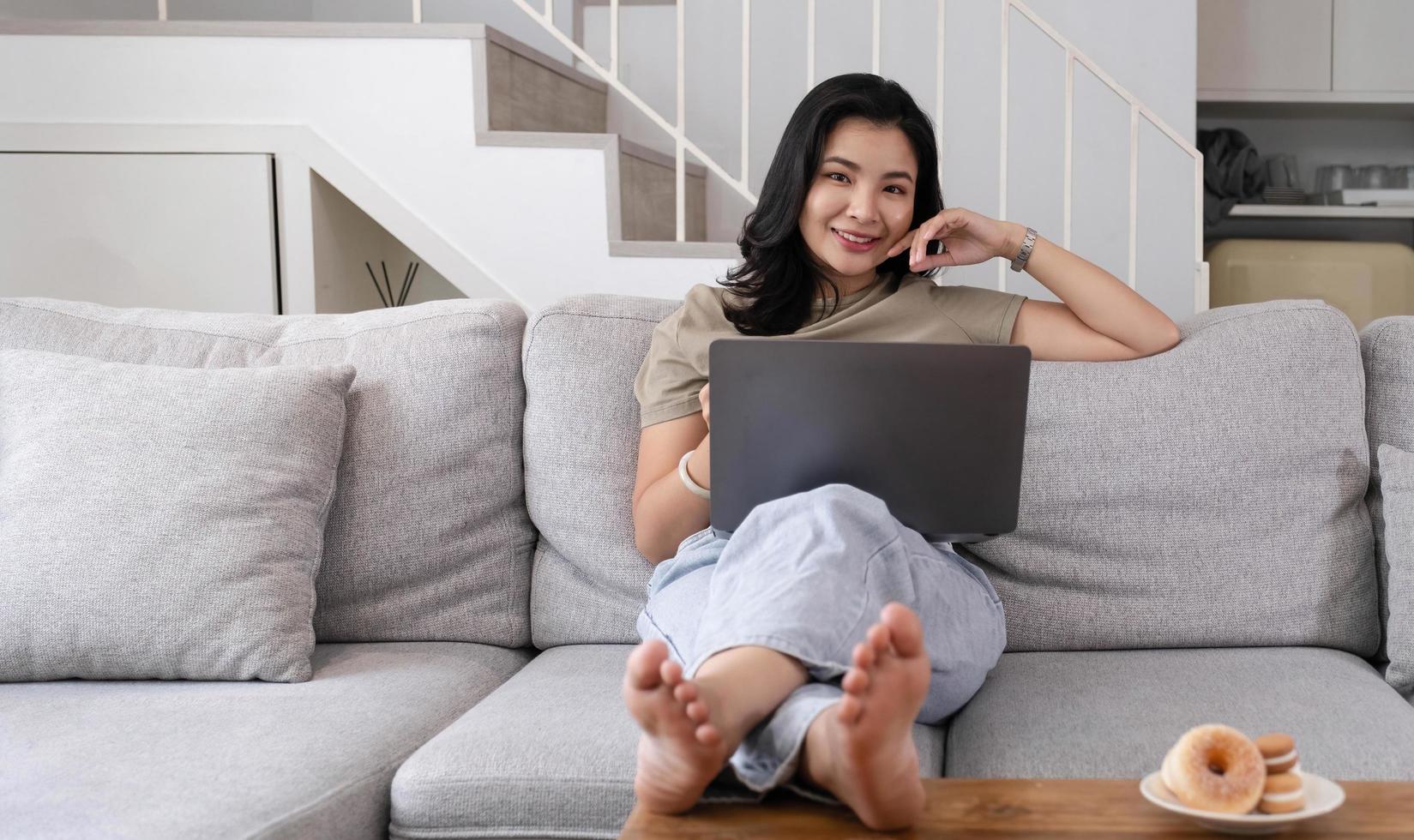 schöne lächelnde asiatische frau, die kamera entspannt auf ledersofa zu hause betrachtet. auf einem Laptop-Computer arbeiten foto