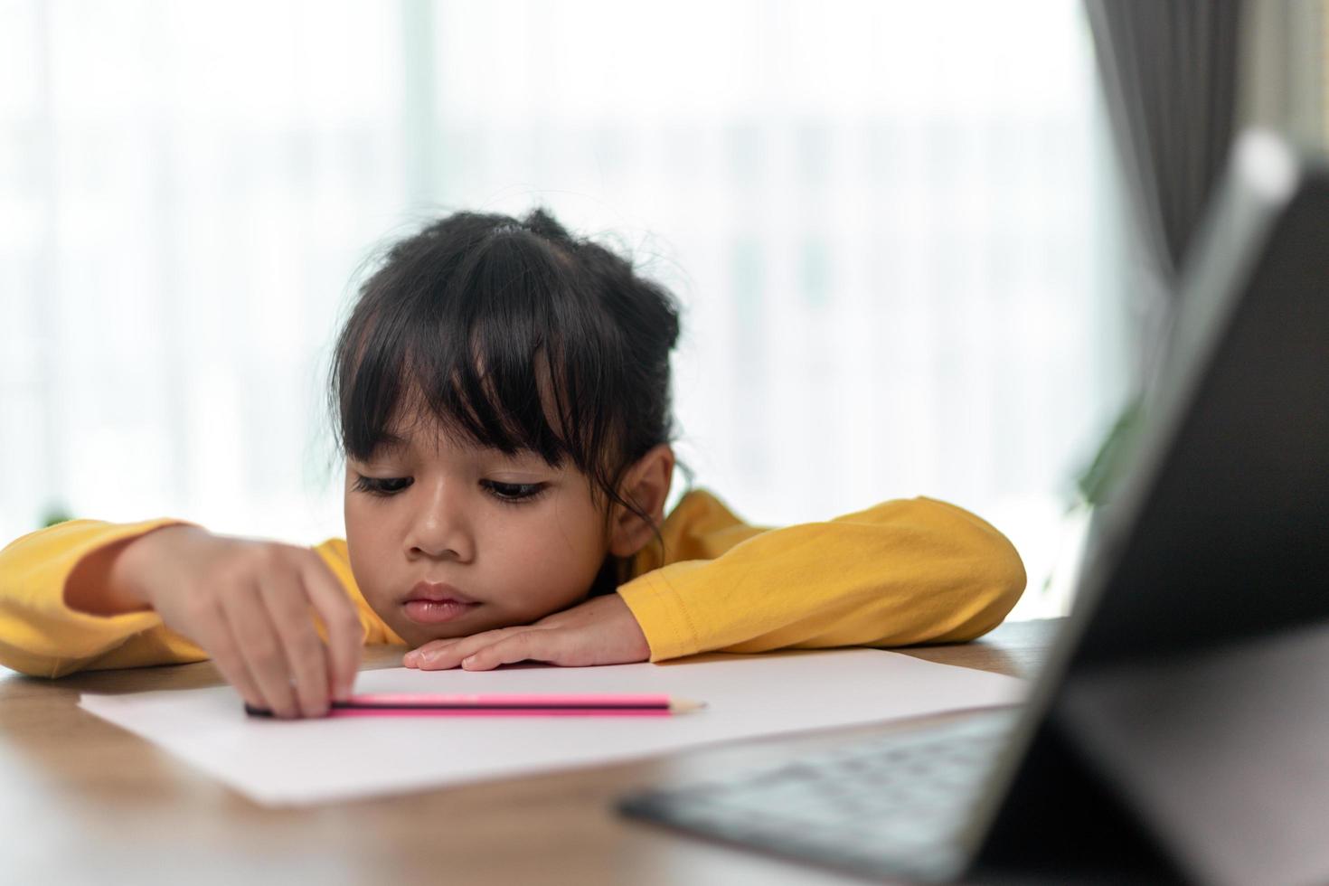 Kleines asiatisches Mädchen, das allein sitzt und mit gelangweiltem Gesicht herausschaut, Vorschulkind, das den Kopf auf den Tisch legt, mit trauriger Langeweile bei den Hausaufgaben, verwöhntes Kind foto