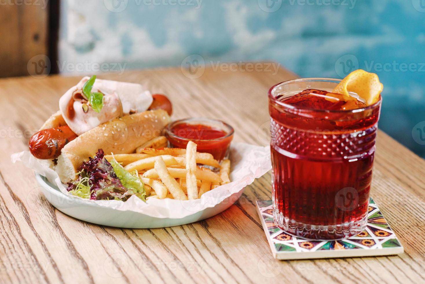 teller mit hot dog mit wurst und schinken, tomatensauce ketchup und französisch friedold fashioned cocktail und auf dem tisch in der bar foto