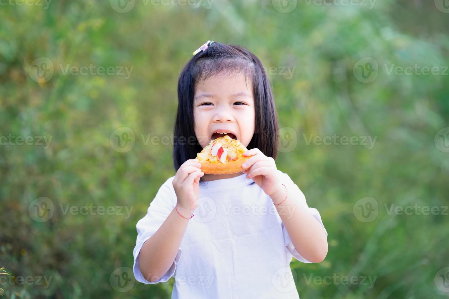 Porträt süßes asiatisches Mädchen isst gerne Pizza mit Gusto. glückliches kind, das essen hält und es mit beißen zu essen in den mund nimmt. Kinder tragen weiße T-Shirts. grüner naturhintergrund. foto