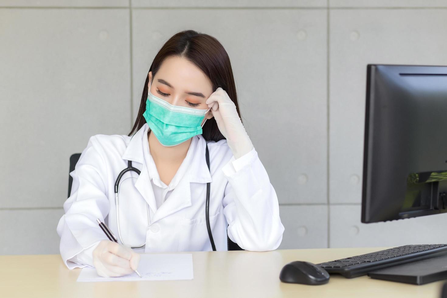 asiatische professionelle ärztin arbeitet ernsthaft, während sie das behandlungsdokument des patienten schreibt. sie trägt eine medizinische maske und latexhandschuhe im klinischen untersuchungsraum des krankenhauses foto