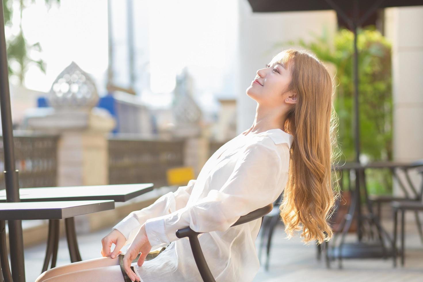 schöne asiatische frau mit bronzefarbenen haaren in einem weißen hemd sitzt glücklich. vor dem Café an einem sonnigen Morgen foto