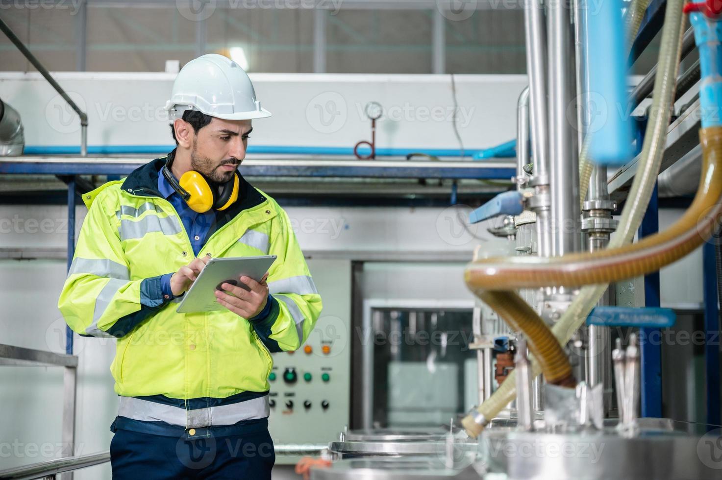 junger männlicher maschineninspektor mit weste und helm mit kopfhörern, der maschine und sterilisatoren in einer wasseranlage überprüft, während er sich notizen in einem digitalen tablet macht foto