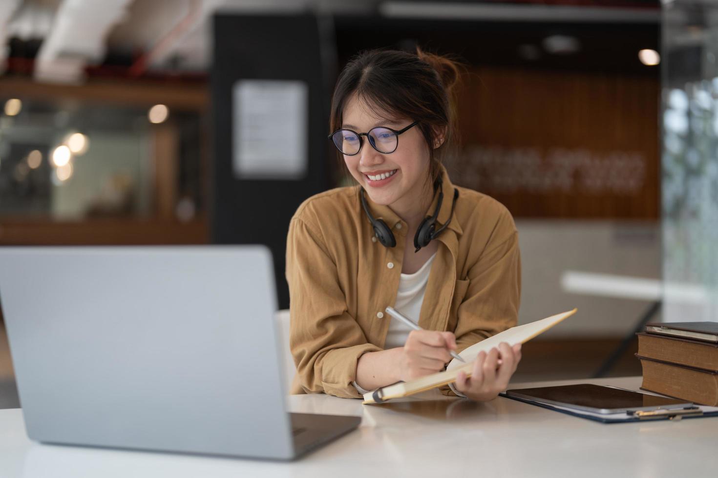 porträt einer jungen asiatischen frau, die einen laptop verwendet und eine liste erstellt, die sich notizen im notizblock macht, die auf dem laptop drinnen arbeiten oder lernen - bildungskurs oder training, seminar, bildungs-online-konzept. foto