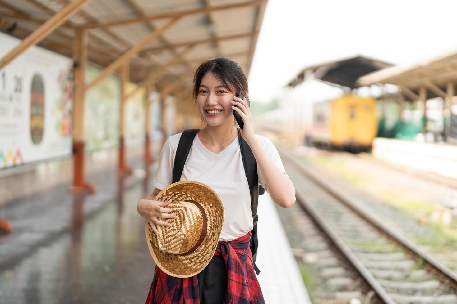 schöne junge asiatin mit einem rucksack benutzt das telefon, während sie in der nähe des eisenbahnzugs auf dem bahnsteig steht. günstiges reisesommerkonzept foto