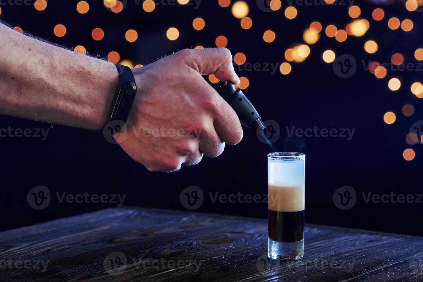 Seitenansicht der Hand des Mannes entzündet Alkohol. Cocktail ist fertig. auf dem Holztisch im Nachtclub. schöne Beleuchtung foto