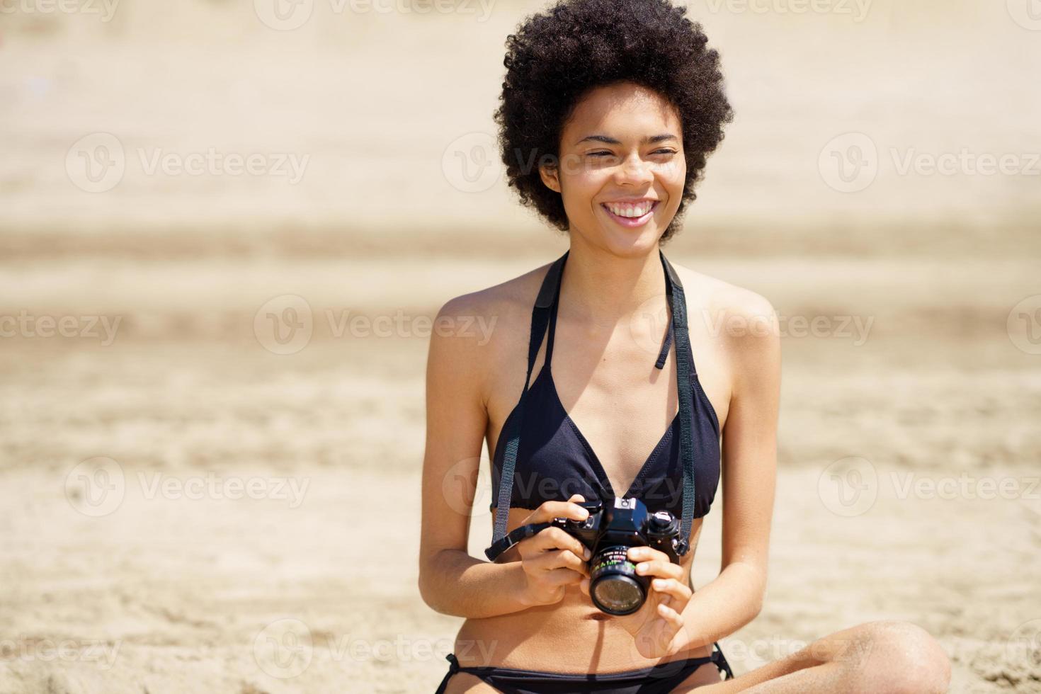 Fröhlicher schwarzer Fotograf in Badebekleidung sitzt am Strand foto