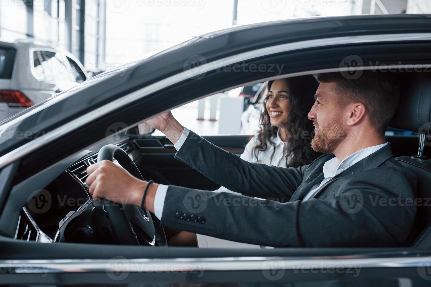 das sieht gut aus. Schönes erfolgreiches Paar, das im Automobilsalon ein neues Auto ausprobiert foto