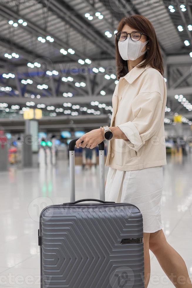 junge frau, die gesichtsmaske mit gepäck trägt, die die flugzeit am flughafen überprüft, infektion mit der schutzkoronavirus-krankheit, reisende asiatin, die bereit ist zu reisen. Neue Normalität und Reisen unter Covid-19 foto