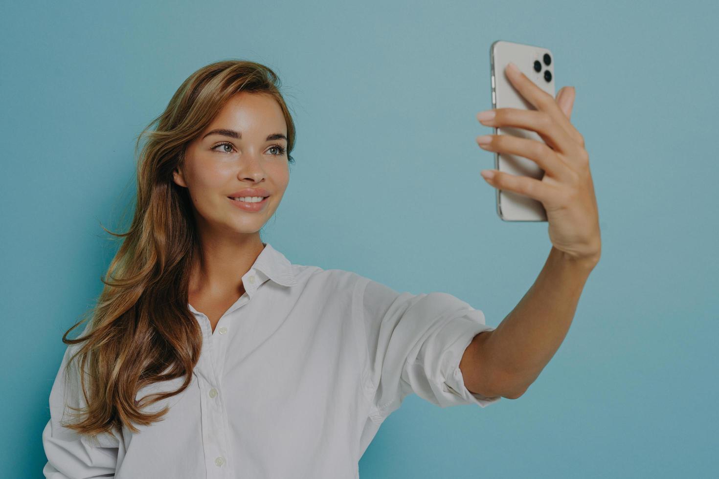 junge elegante europäische frau, die für selfie aufwirft und mit smartphone in der hand gegen blaue wand steht foto