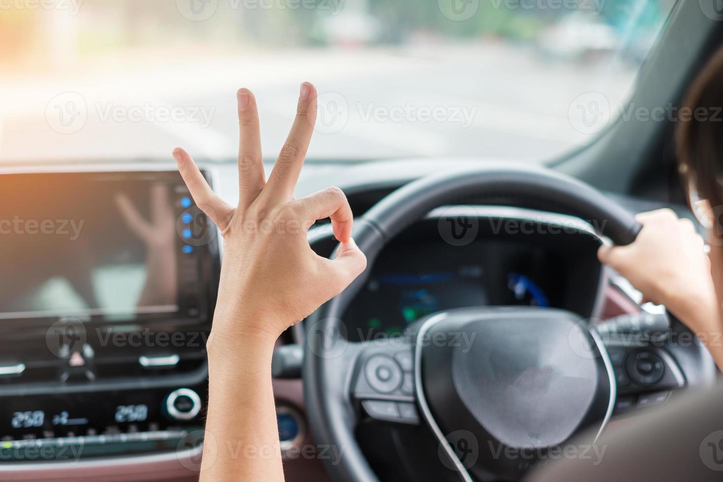 frau, die ok-zeichen zeigt, während sie ein auto auf der straße fährt, handsteuerndes lenkrad in einem elektrischen modernen automobil. Reise-, Reise- und Sicherheitstransportkonzepte foto