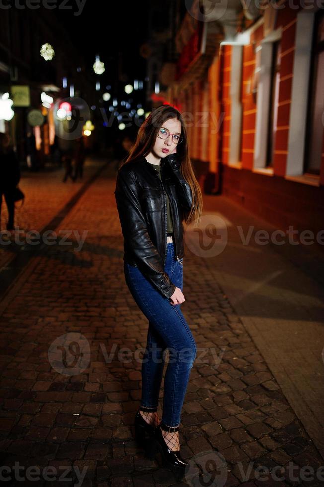 Nachtporträt von Mädchen Model Wear auf Brille, Jeans und Lederjacke, mit Rucksack, gegen die Straßen der Stadt. foto