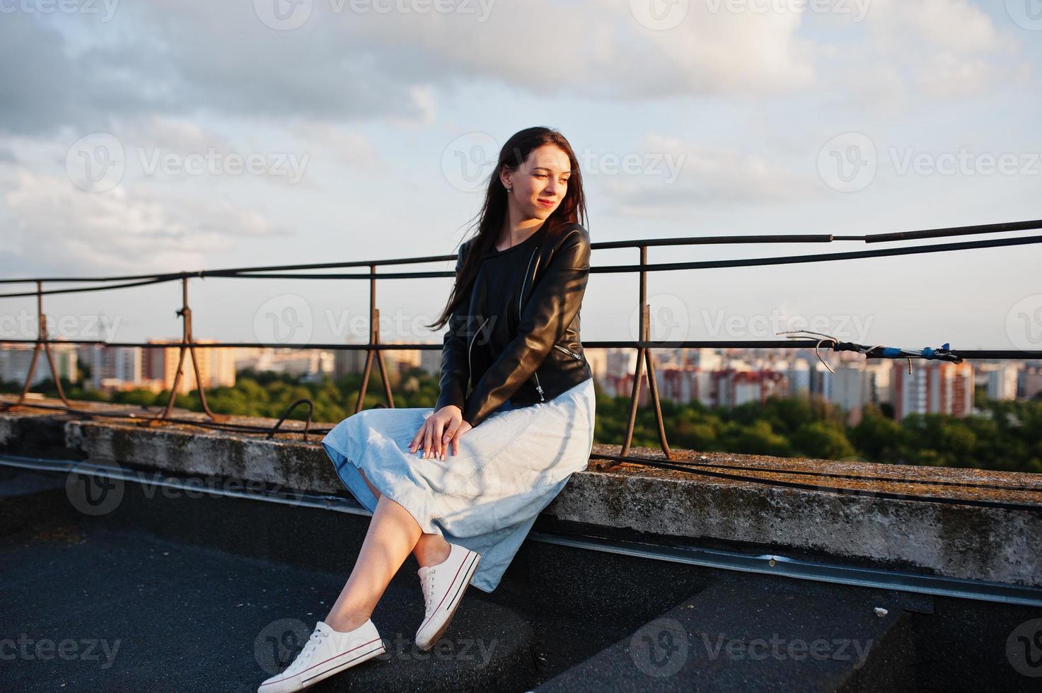 Porträt eines hübschen Mädchens in blauem Kleid, schwarzer Lederjacke und weißen Turnschuhen, das auf dem Dach sitzt und einen herrlichen Blick auf eine Stadt bietet. foto