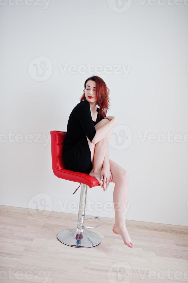 Rothaariges Mädchen in schwarzer Kleidertunika sitzt auf rotem Stuhl an weißer Wand im leeren Raum. foto