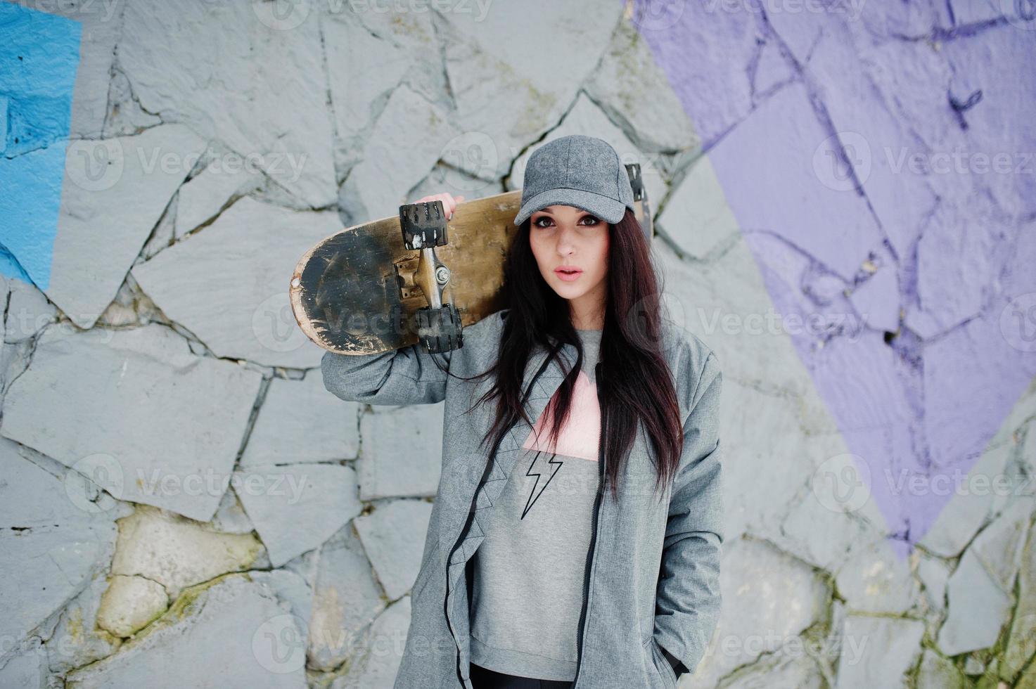 stilvolles brünettes mädchen in grauer kappe, lässiger streetstyle mit skateboard am wintertag gegen farbige wand. foto