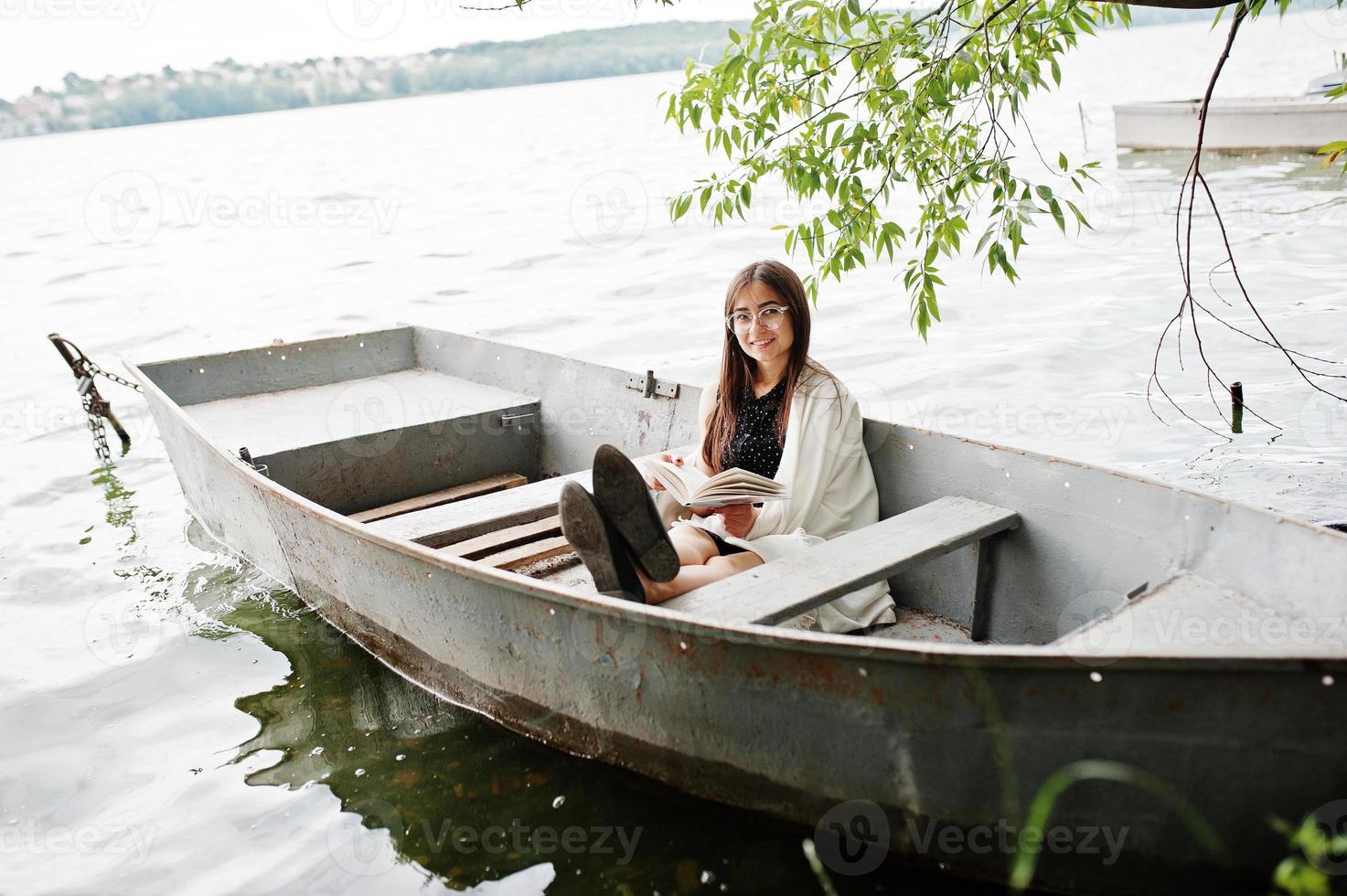 Porträt einer attraktiven Frau mit schwarzem Tupfenkleid, weißem Schal und Brille, die in einem Boot auf einem See ein Buch liest. foto