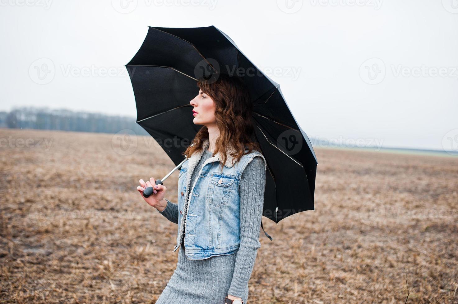 Porträt eines brünetten lockigen Mädchens in Jeansjacke mit schwarzem Regenschirm auf dem Feld. foto