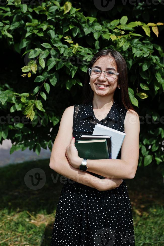 Porträt einer schüchternen jungen Frau in schwarzem Tupfenkleid und Brille, die Bücher im Park hält. foto