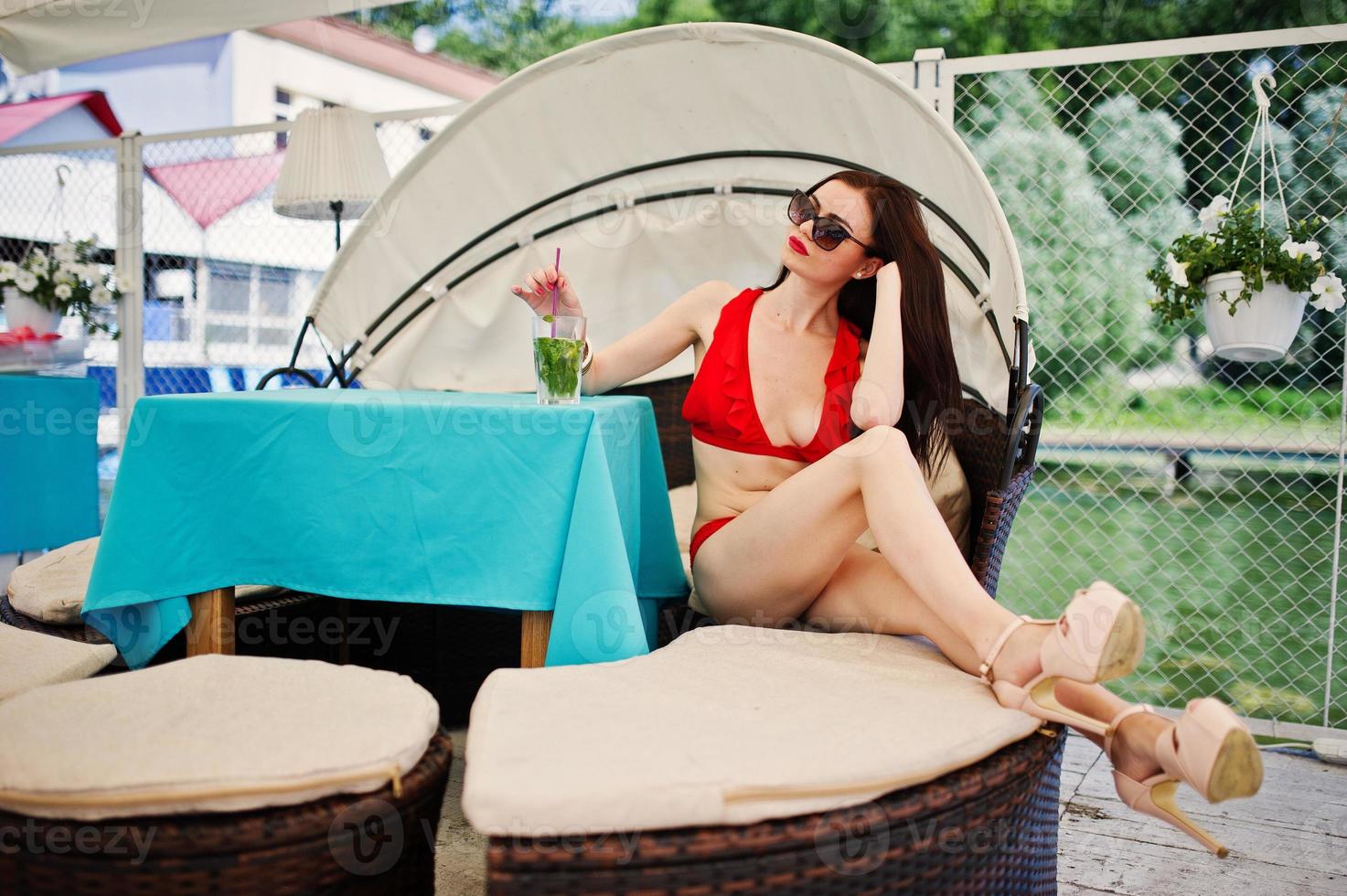 Porträt eines atemberaubenden Mädchens in rotem Bikini-Badeanzug und Sonnenbrille, das ihre Zeit genießt, indem es auf dem Sofa am Kai am See sitzt. foto