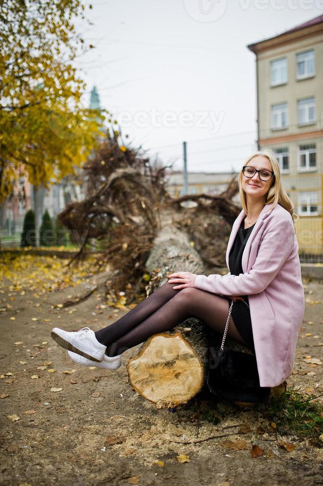 Blondes Mädchen mit Brille und rosafarbenem Mantel, schwarze Tunika, die auf gefälltem Baum sitzt. foto